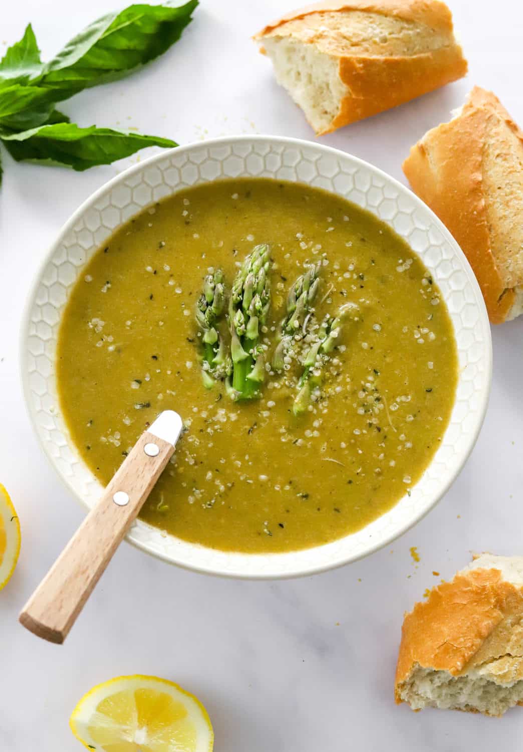 Vibrant green blended soup in a white bowl topped with asparagus ends with broken bread and herbs scattered around it. 