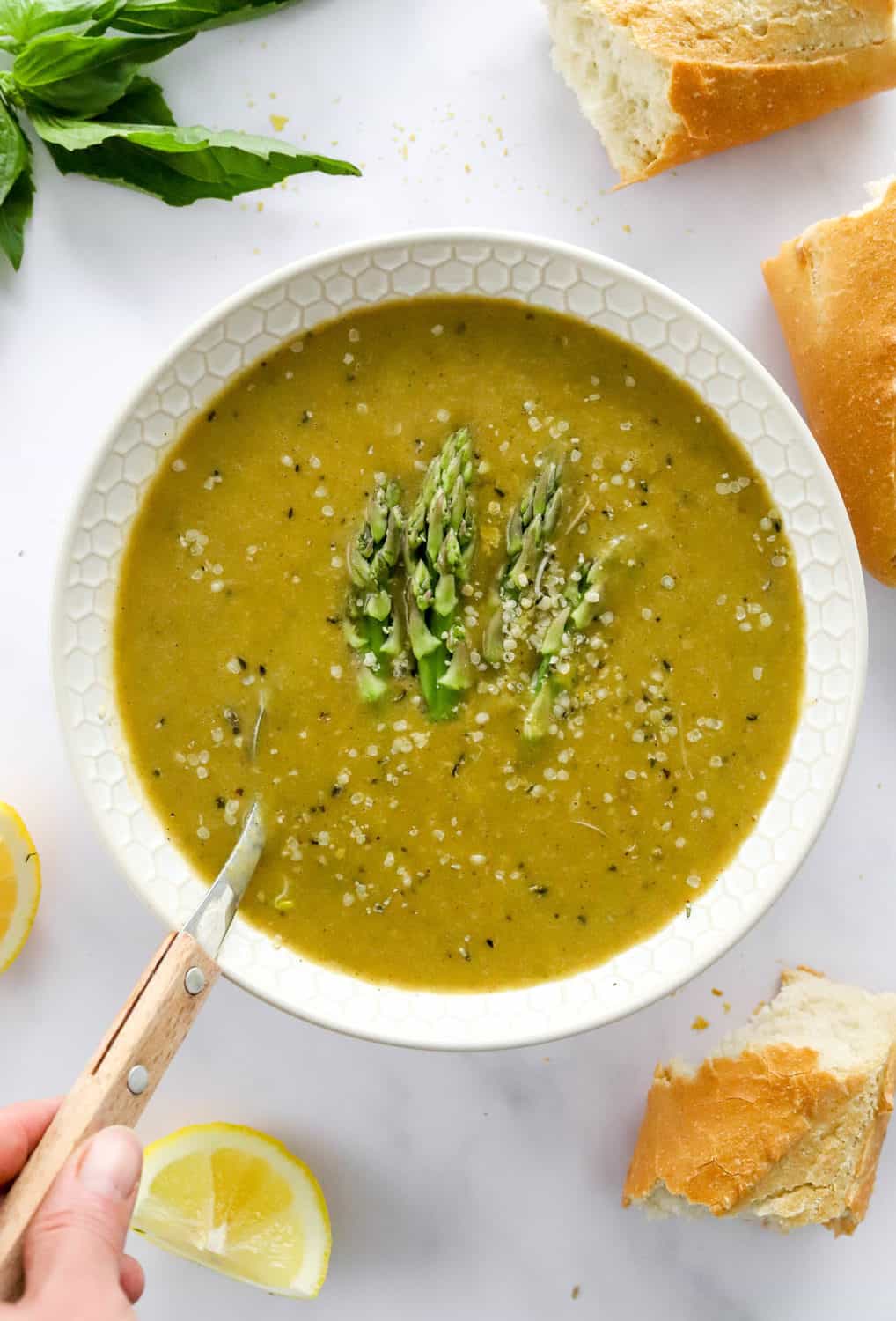 Vegan asparagus soup in a white bowl with a spoon in it with torn bread and basil around it. 