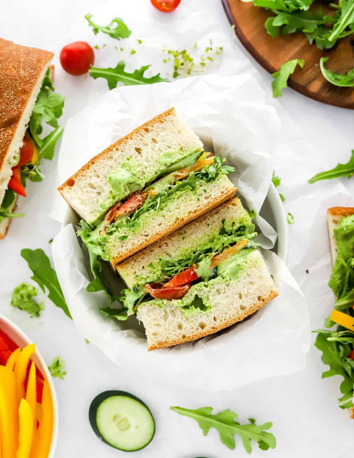 Vegan sandwich loaded with veggies and a creamy green dressing in between large slices of bread in a bowl with more sandwiches around it. 
