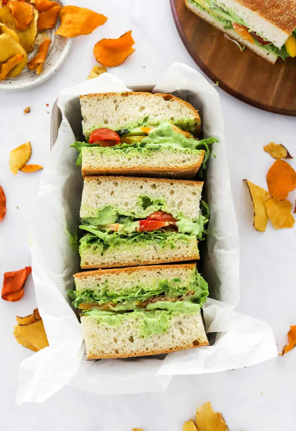3 veggie vegan Sandwiches in a rectangle tin on top of white paper with veggies chips around it. 