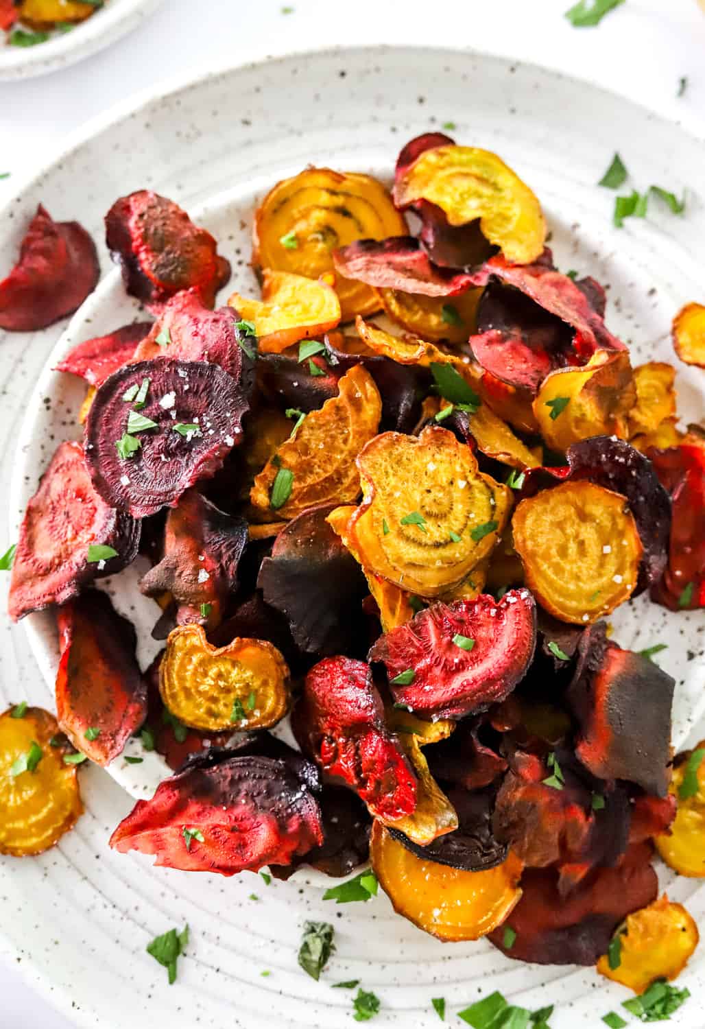 Crispy baked beet chips on a white plate with green herbs on top with another plate of beet chips behind it. 