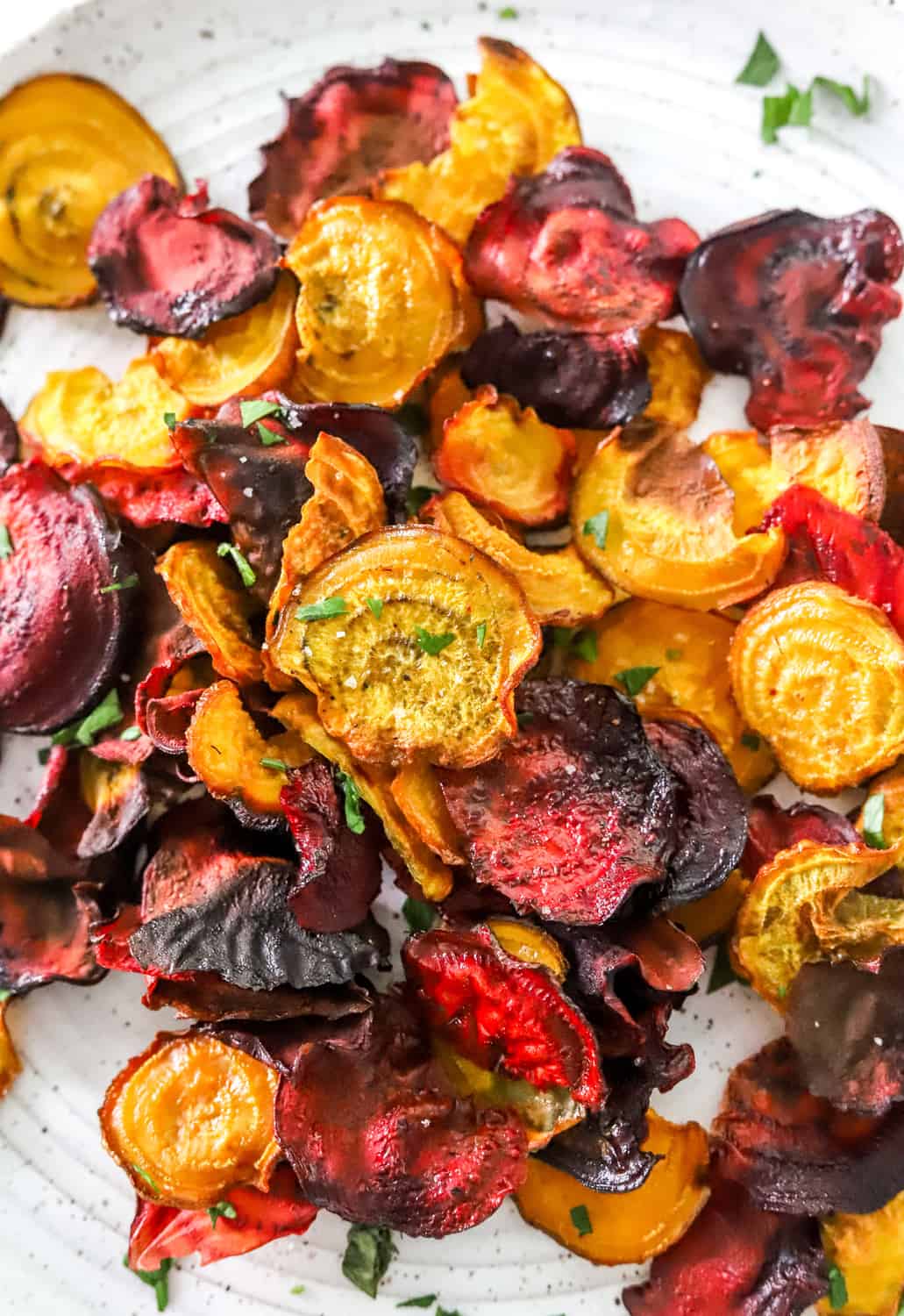 Golden and red round veggie chips spread out on a white plate with sea salt and chopped herbs sprinkled on top of them. 