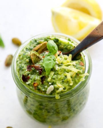 Round jar filled with green pesto topped with lemon zest and nuts with more sliced lemon and nuts behind it