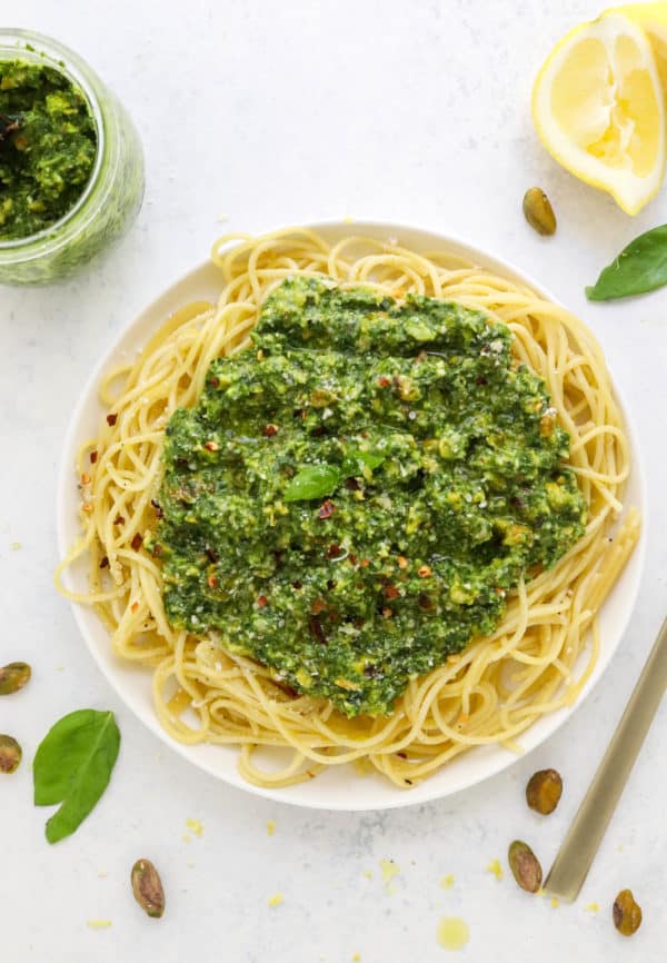Round white bowl of spaghetti with a pile of green pesto sauce on top if it with a jar of pesto and sliced lemons behind it on a white surface