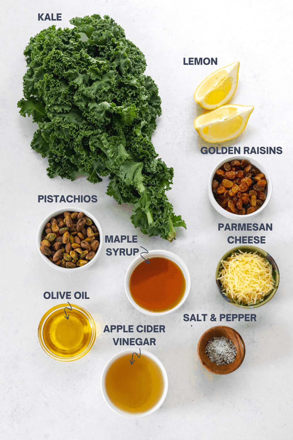 Kale, lemon, bowl of nuts, bowl of raisins, oil and vinegar on a white surface with labels over the top of each ingredient. 