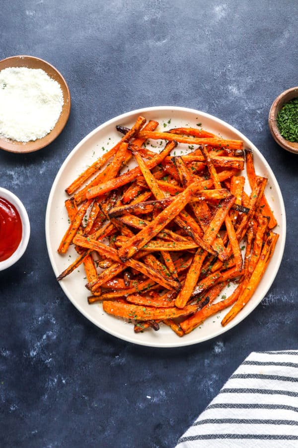 Round beige plate with a pile of golden brown carrot fries on it topped with herbs and shredded parmesan cheese with a small bowl of ketchup next to it