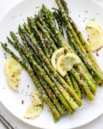 Cooked asparagus in a pile on a round white plate with sliced lemon laying on top of it