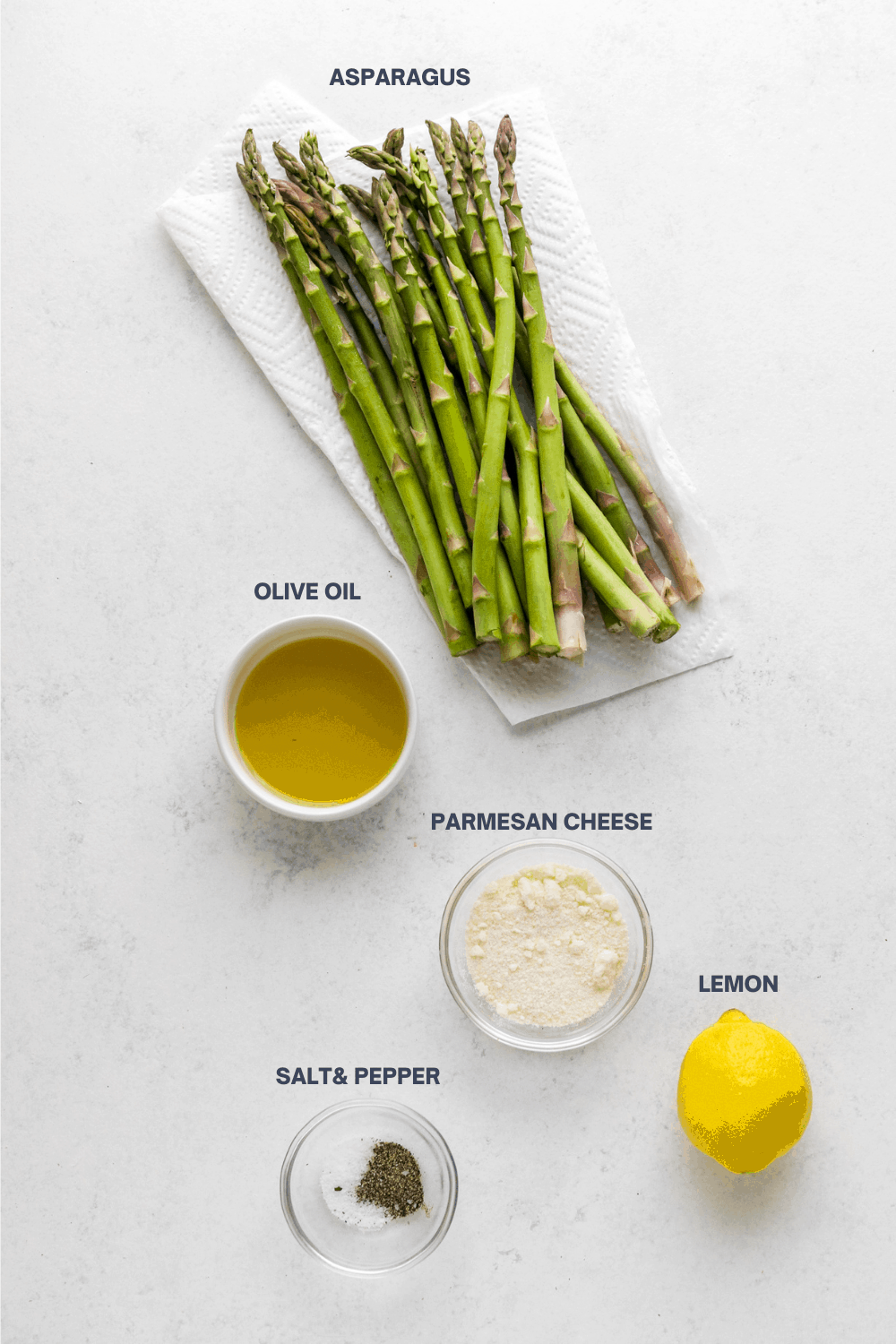 Raw asparagus, olive oil in a white bowl, shredded parmesan cheese, lemon, salt and pepper on a white surface with labels above them. 