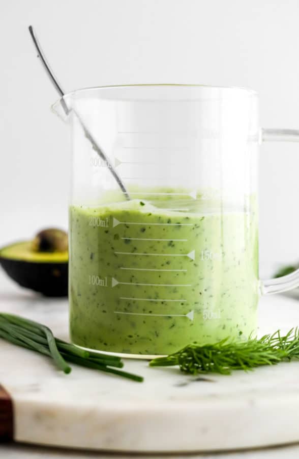 Tall glass measuring cup filled with cray green salad dressing with a spoon in it with herbs around it and part of a sliced avocado behind it