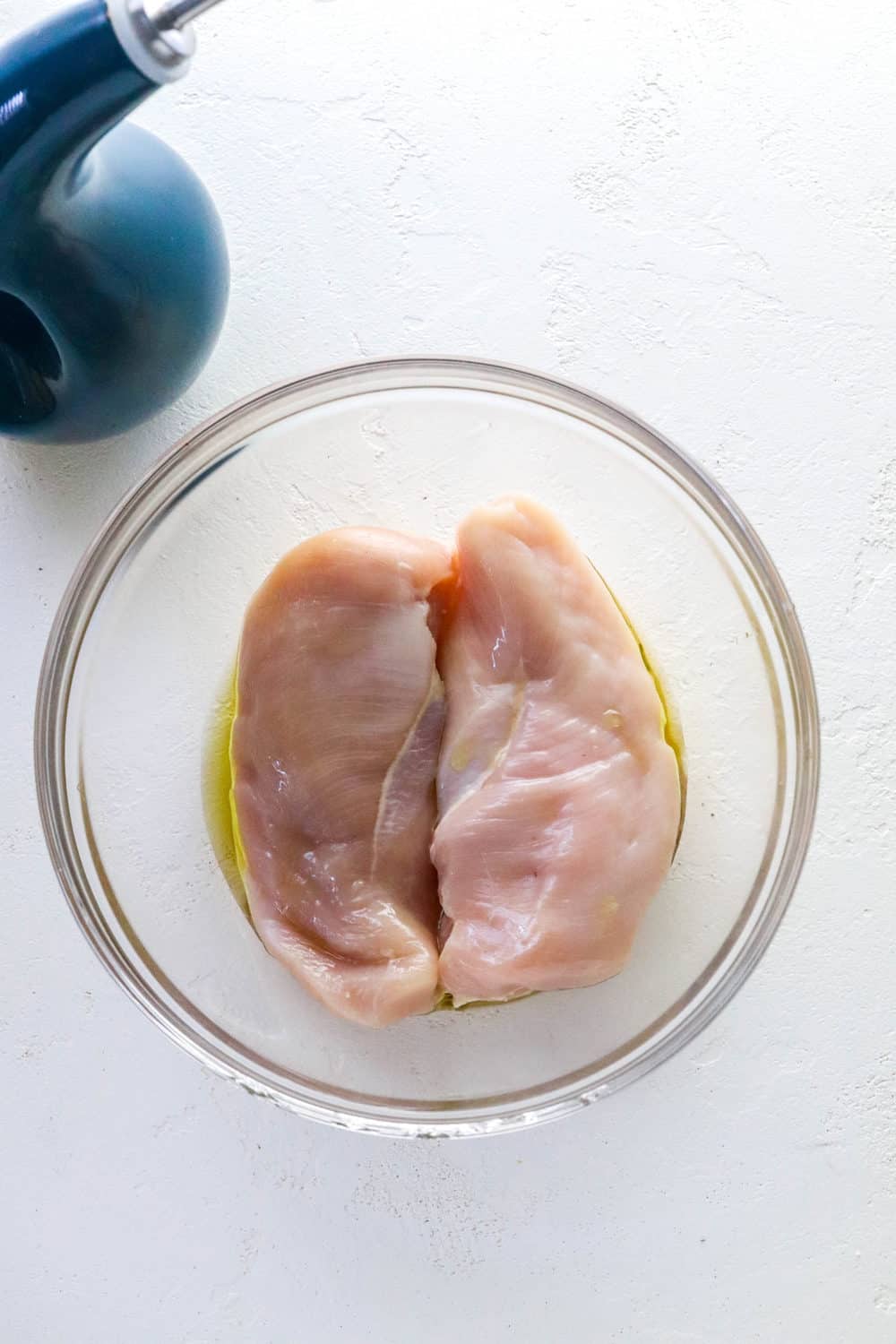 Two pieces of raw boneless chicken breast in a round glass bowl with olive oil on them