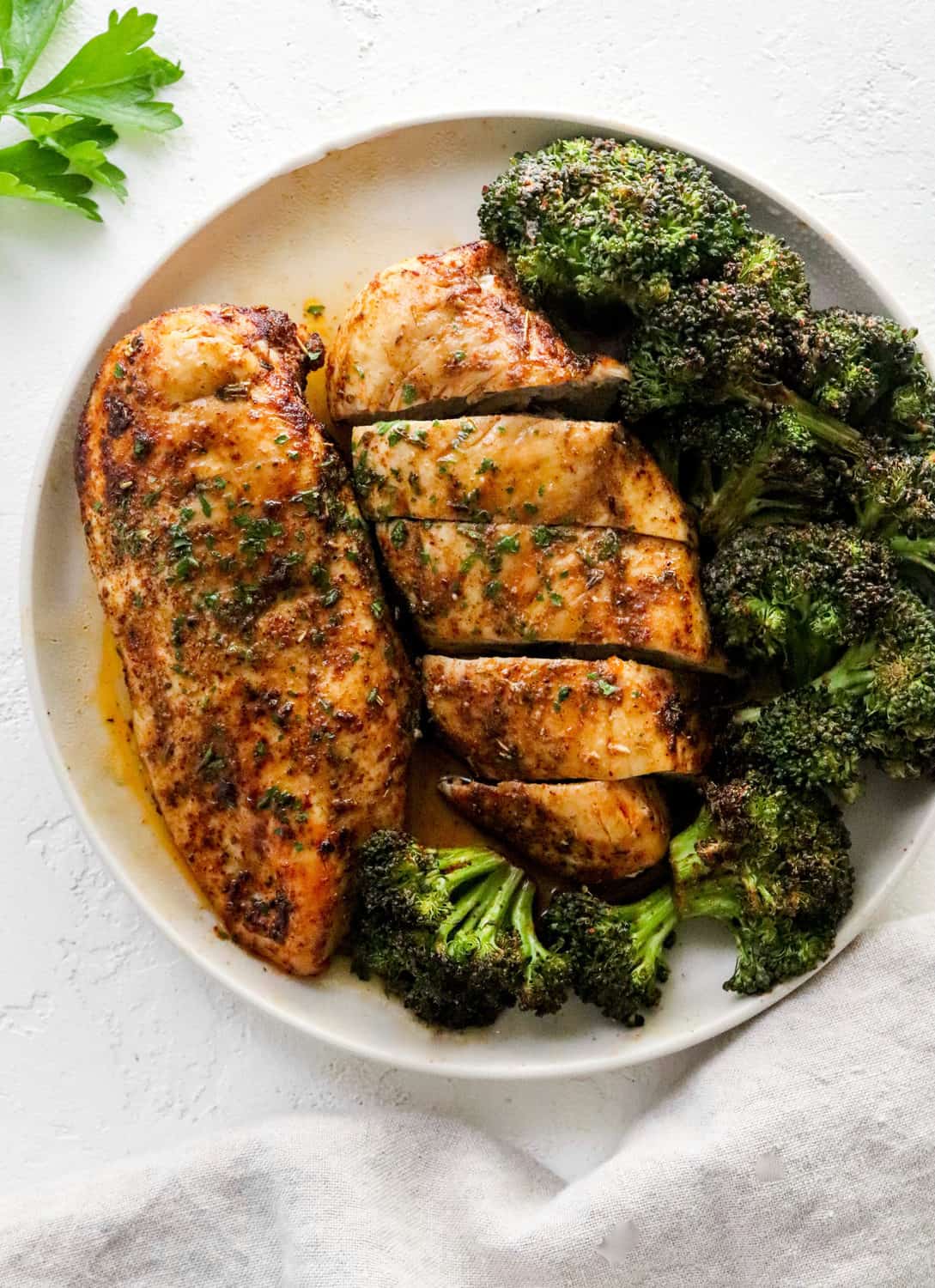 Seasoned cooked chicken beast on a round white plate with another pieces of sliced chicken next to it and cooked broccoli around it on the plate