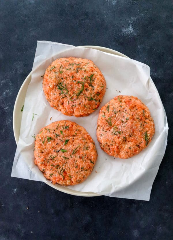 3 raw formed salmon patties on  parchment paper on a round plate on top of a dark blue surface