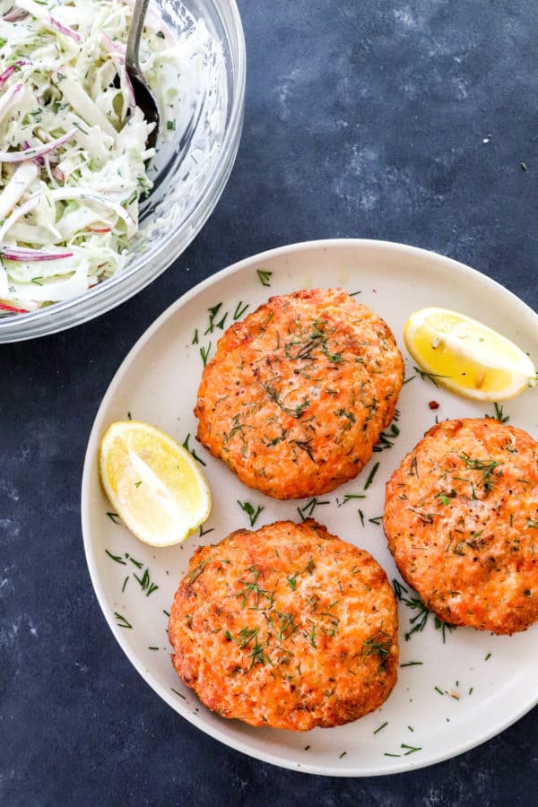 3 cooked salmon patties on a round plate with dill on them and lemon wedges on the plate with a bowl of creamy Cole slaw behind it