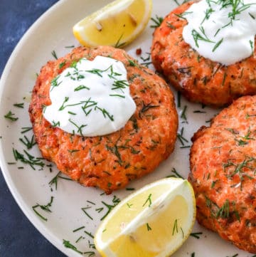 3 air fryer salmon patties on a plate topped with greek yogurt with lemon wedges on the plate next to them.