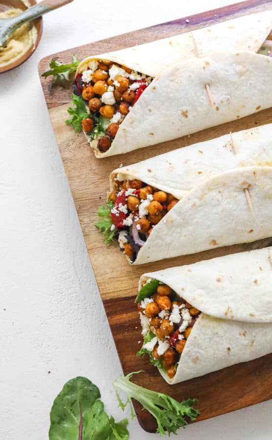 A wooden board topped with three wrapped white flour tortillas filled with crispy roasted chickpeas and veggies and topped with crumble white cheese with a bowl of hummus behind it.
