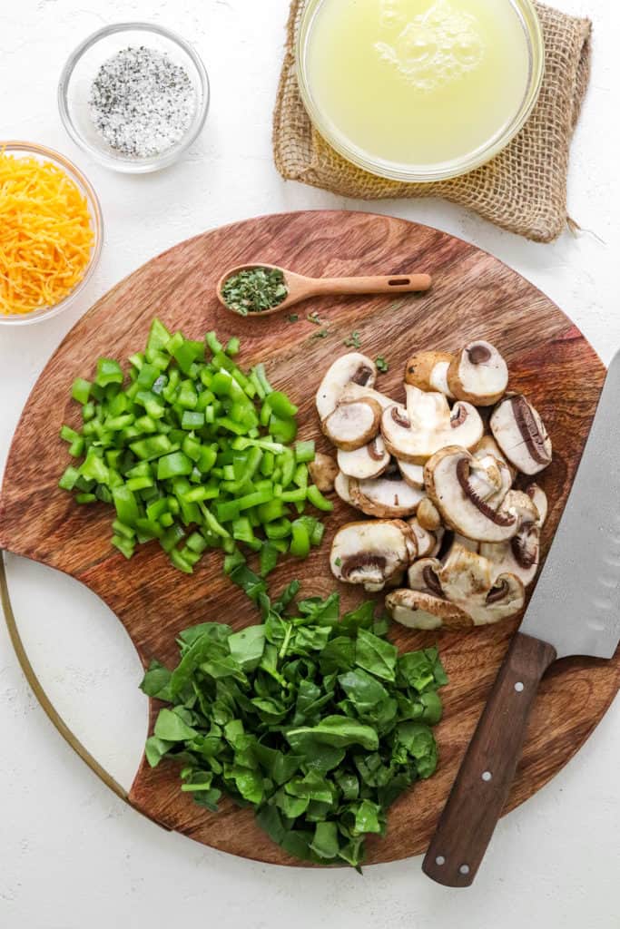 Round wooden cutting board with sliced mushrooms, chopped green peppers and chopped spinach on it with a knife on the edge of the board. 