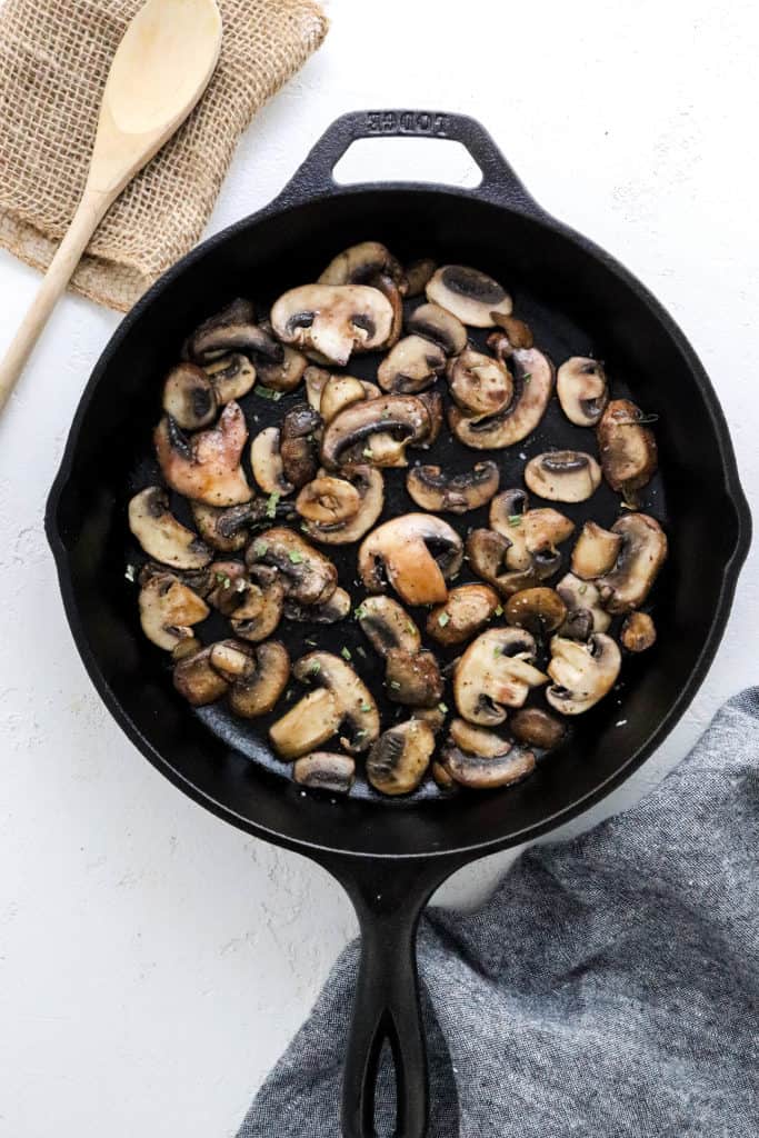 Cast iron pan filled with sliced cooked mushrooms with a wooden spoon behind it and a grey cloth in front of it. 