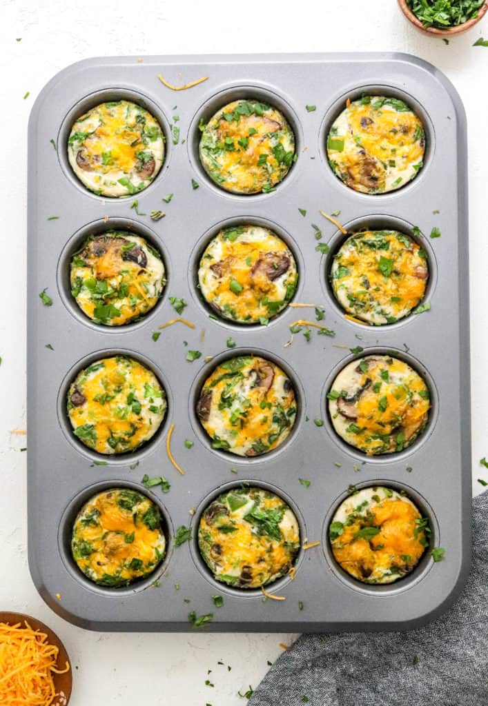 Baked egg white muffins with green and yellow in them in a grey muffin pan with herbs sprinkled on top of them