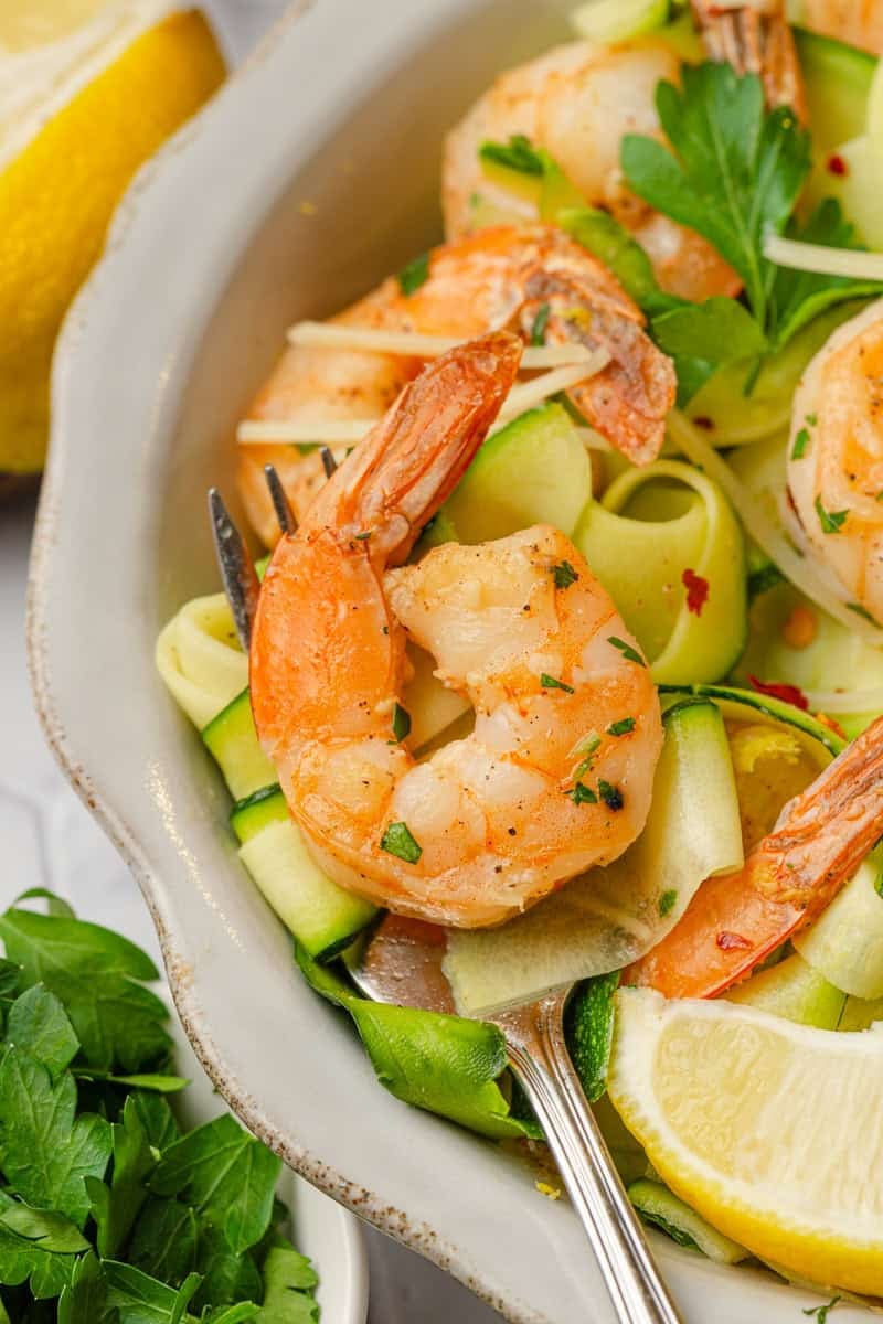 Shrimp and zucchini cooked in a bowl with some on a fork with a lemon slice next to it. 