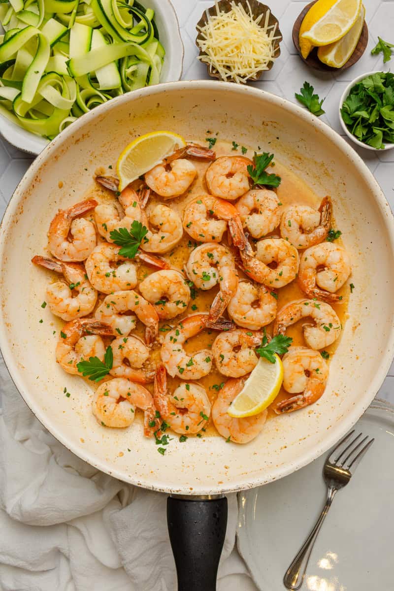 Shrimp scampi cooked in a pan with a bowl of zucchini noodles behind it. 
