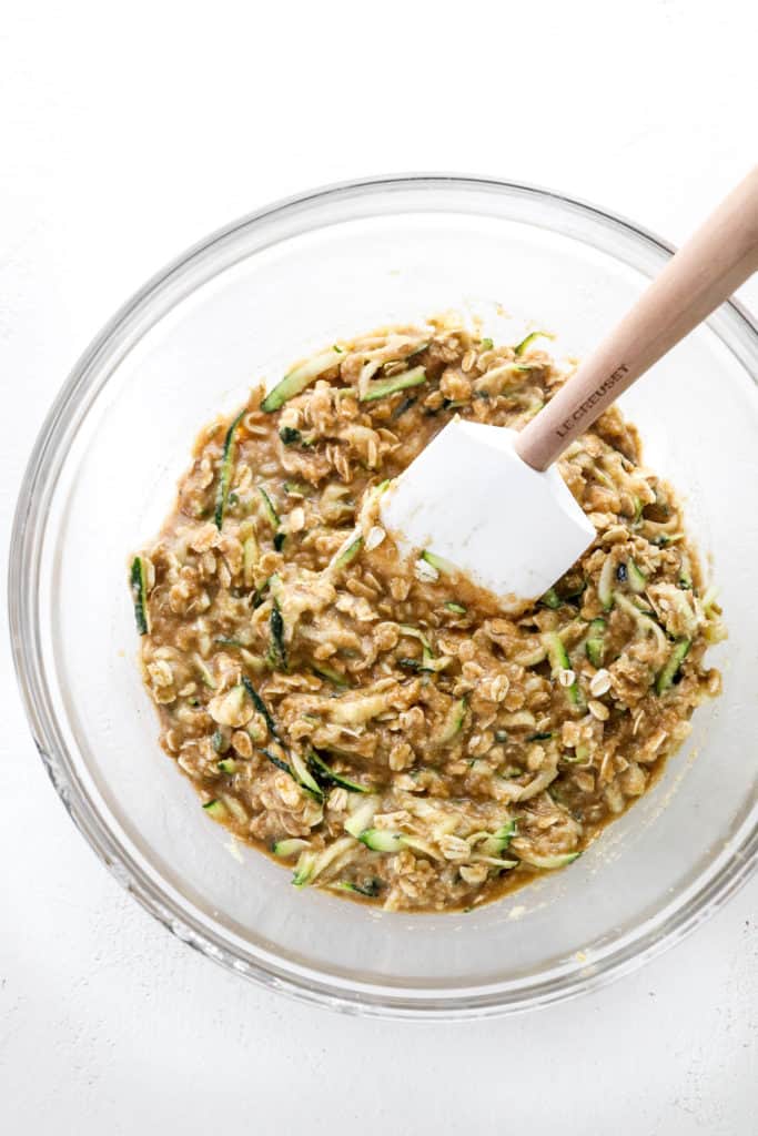 Healthy zucchini muffin batter mixed up in  glass mixing bowl with a white rubber spatula in the batter. 