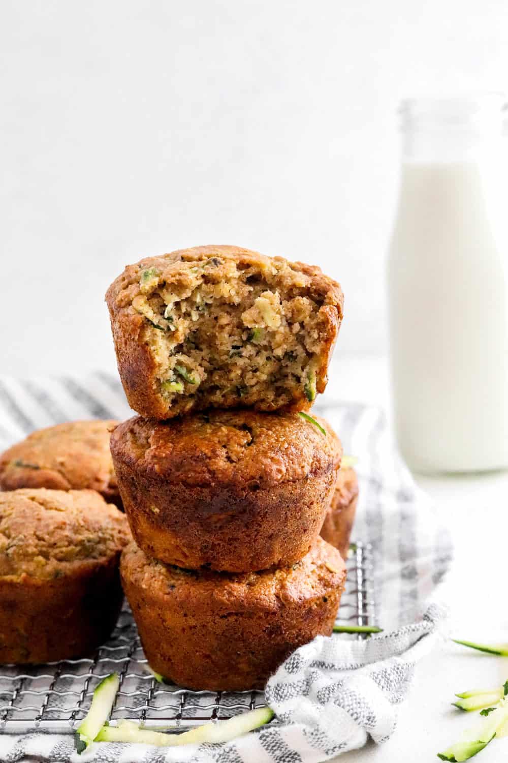 Stack of 3 muffins with a bite taken out of the one on the top with more muffins next to it sitting on a wire rack with a towel with stripes under it and glass of milk behind the muffins. 