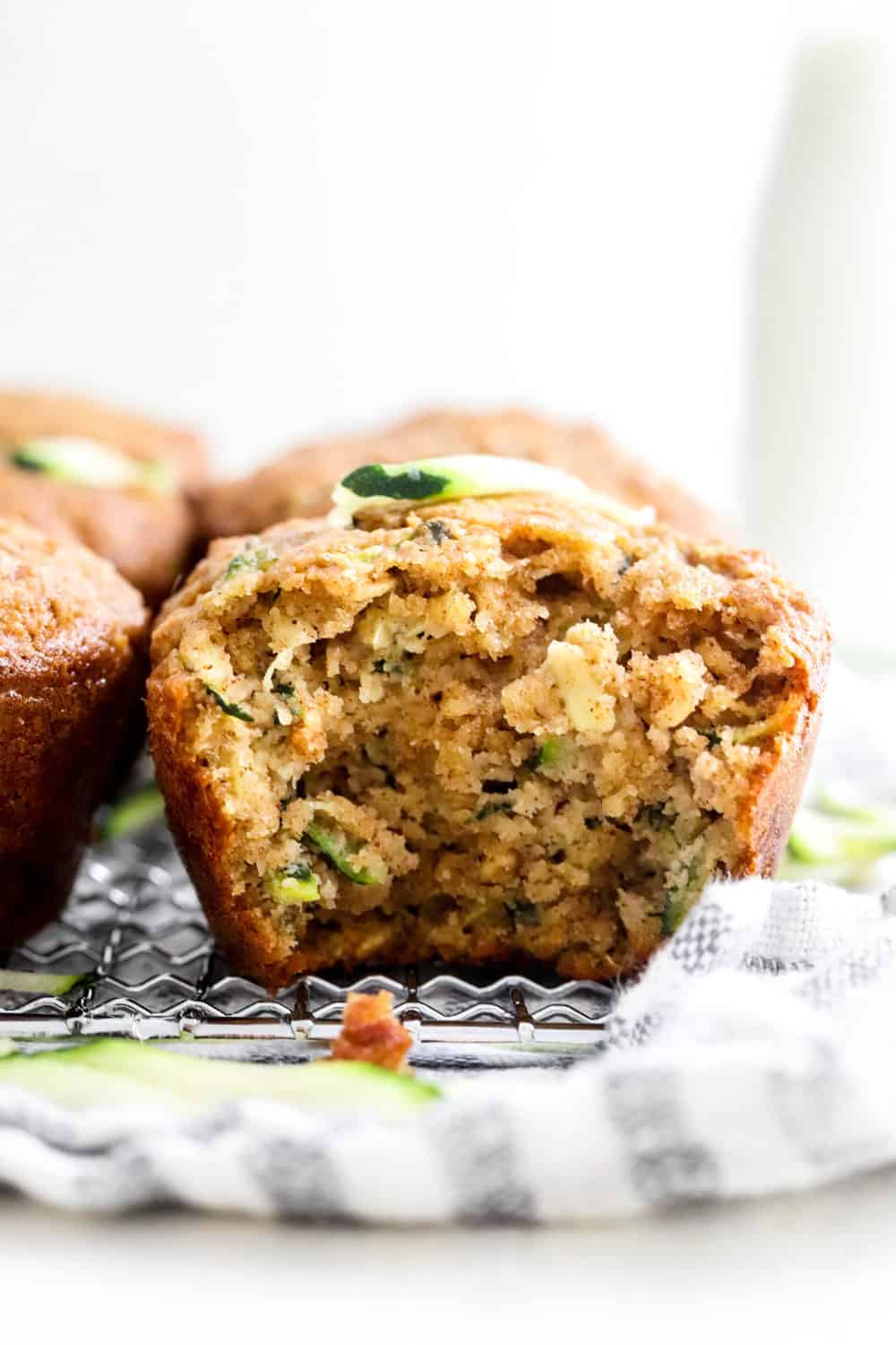 Close up of a gluten free zucchini muffin with a bite taken out of it with more muffins next to it sitting on top of a wire rack with a striped kitchen towel underneath it. 