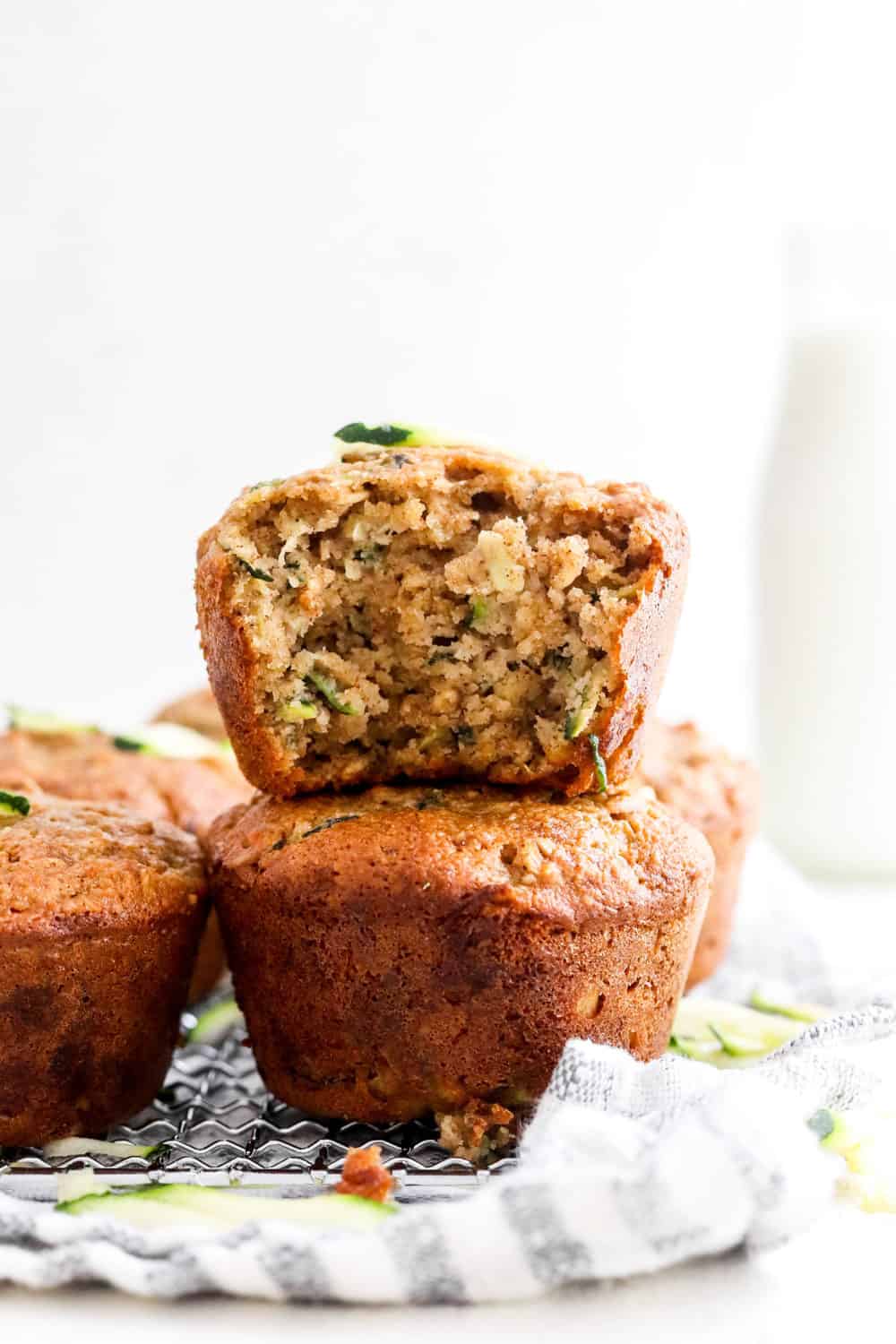 Brown muffin with specs of zucchini in it with a bite taken out of it on top of another whole muffin with more baked muffins next to it and begin it. 