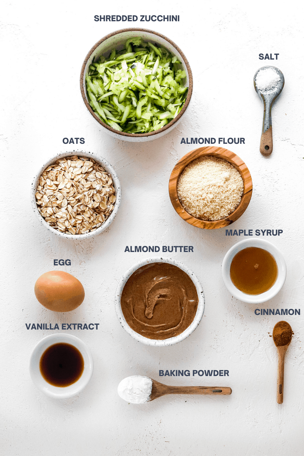 Zucchini muffin recipe ingredients in bowls on a white surface with labels over the top of each ingredient. 