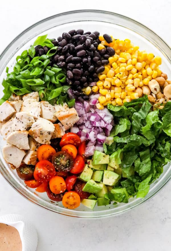 Large round glass bowl with lettuce, diced chicken, corn, black beans sliced tomatoes, red onion and diced avocado separated in the bowl.