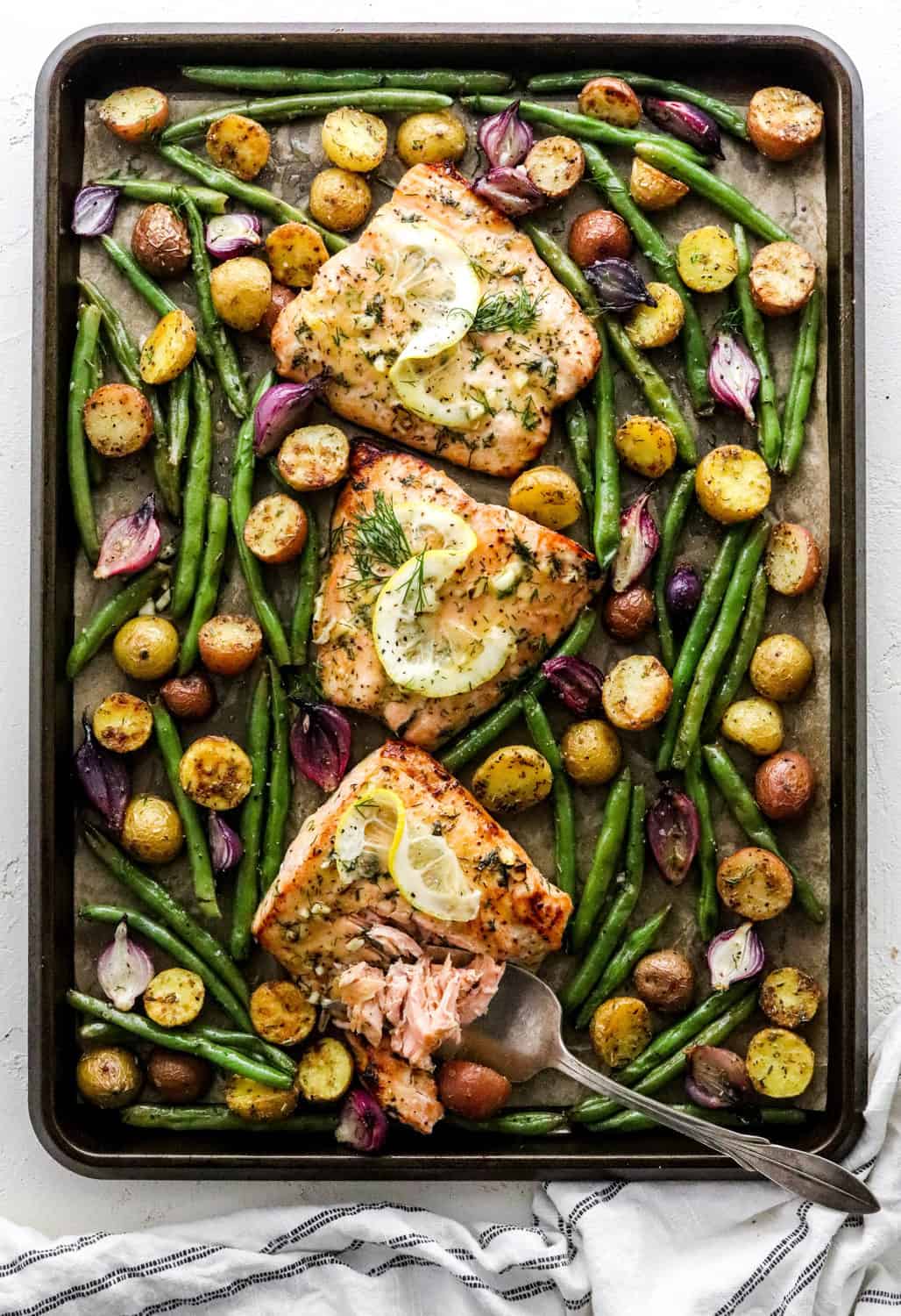 Cooked salmon filets, surrounded by cooked green beans, purple baby onion and baby potatoes spread around the salmon on a sheet pan
