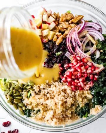 Pouring yellow dressing over a kale and quinoa salad with pomegranate seeds and sliced red onion on it.