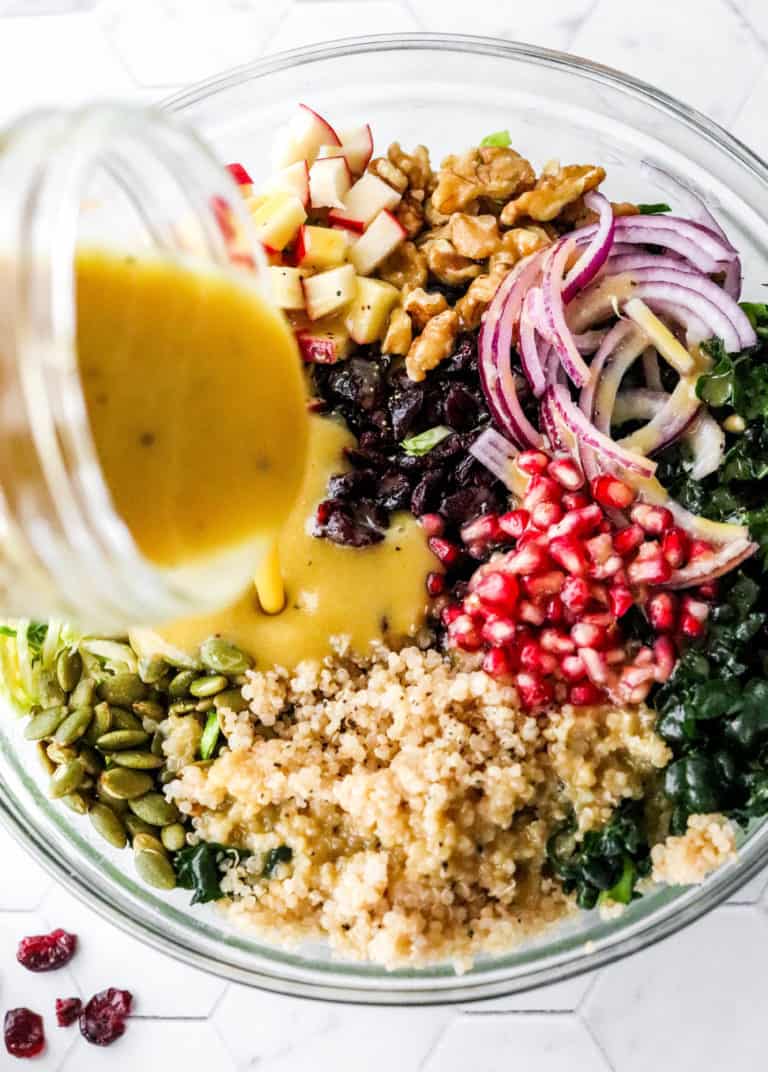 Mustard dressing being poured over a quinoa and kale chopped salad with pomegranate seeds, sliced red onion and nuts on the salad. 