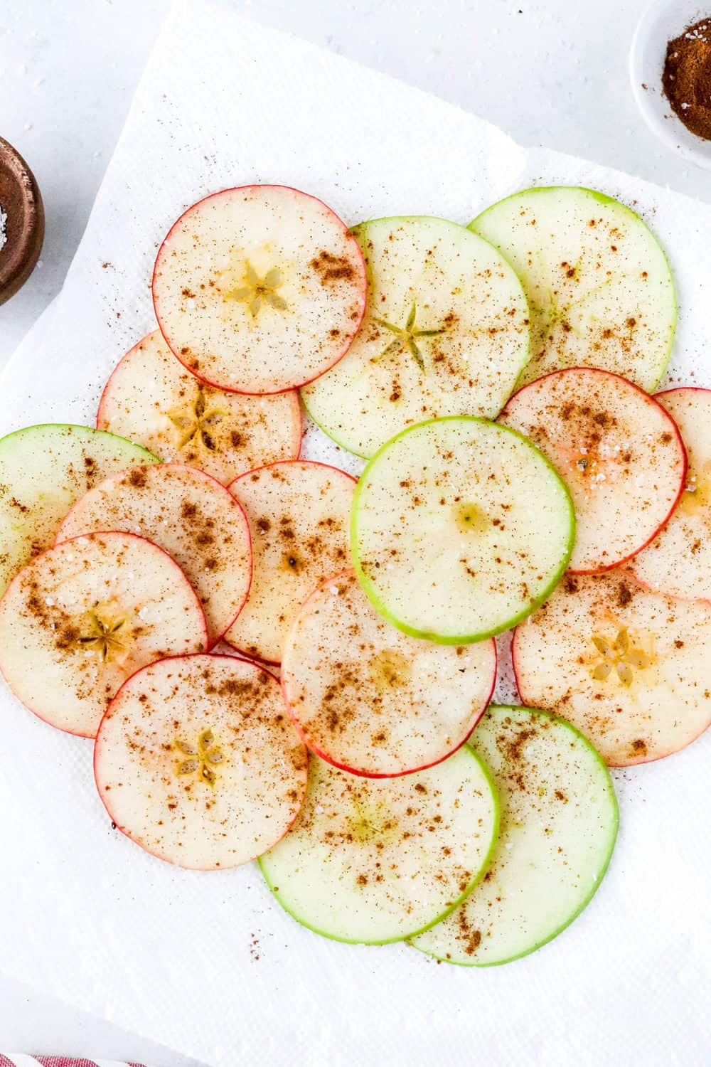 Pile of sliced apples on a white plate sprinkled with cinnamon with a small bowl of cinnamon behind it