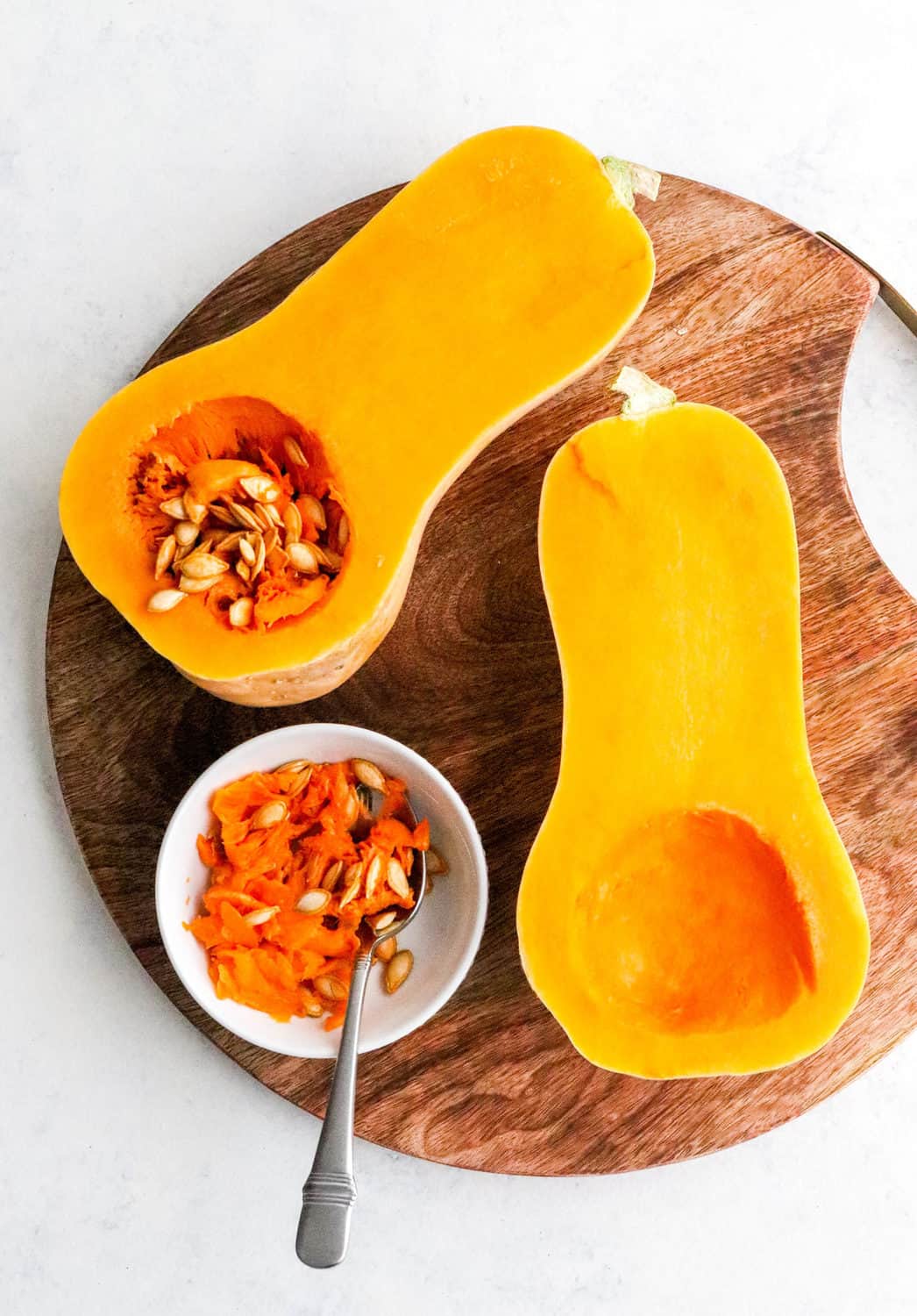 Two halves of butternut squash on a round wooden board on a white surface with a round white bowl with seeds of of the squash and a spoon in it on the board next to the squash.