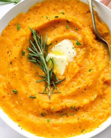 Round white serving bowl filled with mashed butternut squash with a piece pf rosemary in the middle of it