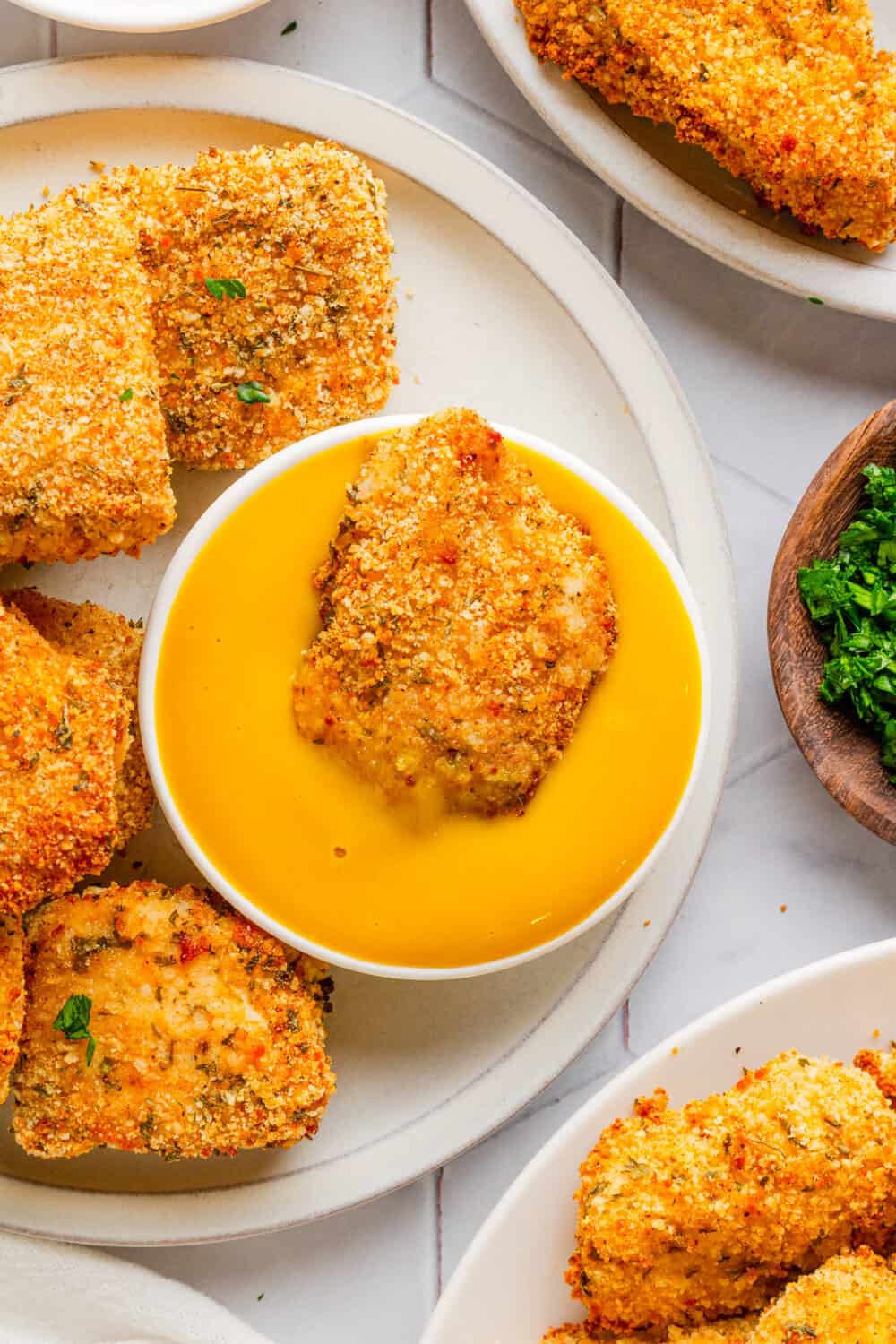 Crispy chicken nugget in a bowl of yellow mustard sauce with more on the plate with it. 