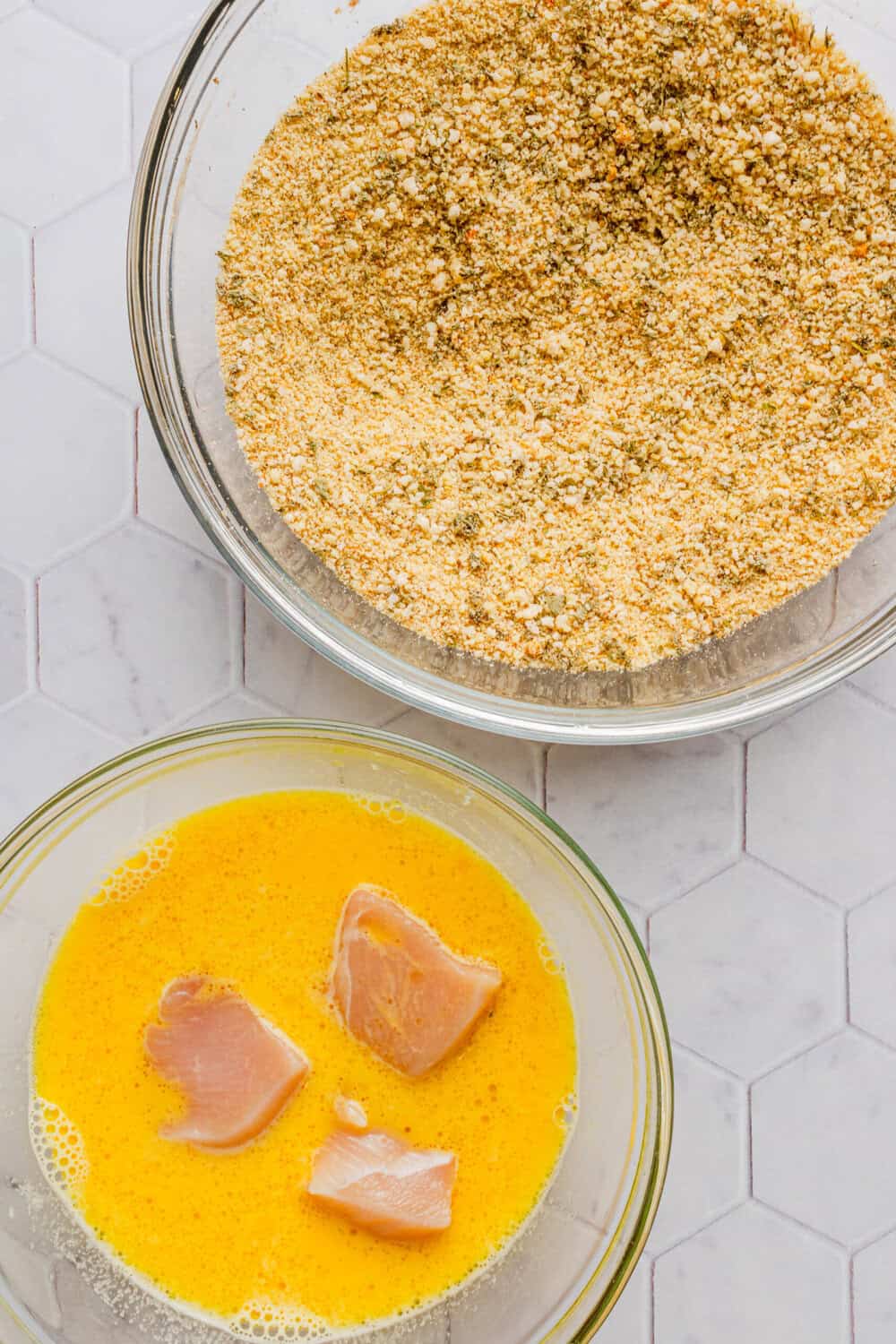 Bowl of parmesan breadcrumbs with a bowl of whisked eggs with three chunks of raw cubed chicken breast in the bowl with the eggs. 