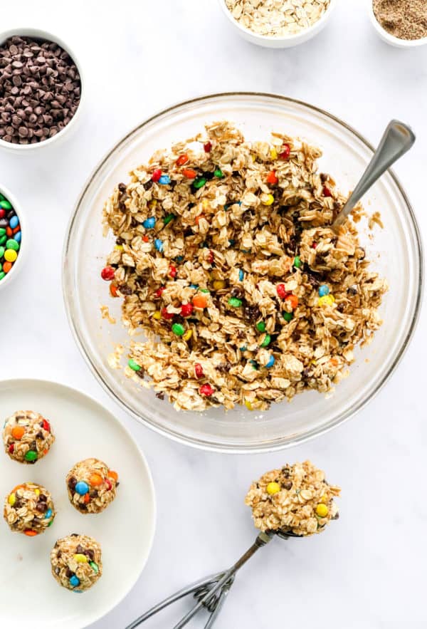 Healthy, chunky energy bite ingredients mixed up into a chunky cookie-like batter in a glass mixing bowl with a plate of rolled energy balls and a scoop of the mixture in front of it.
