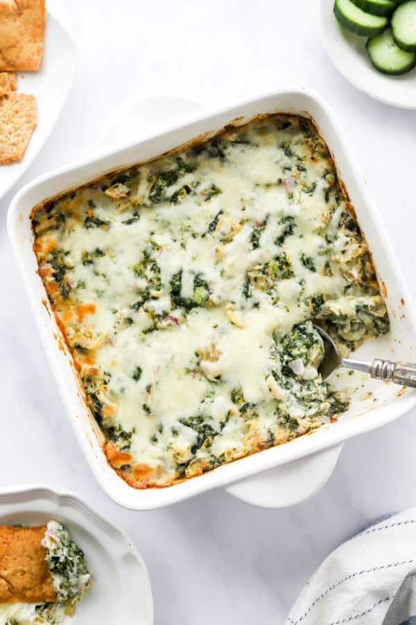 Baked cheesy dip with greens in a white dish with a spoon in tit with chips on a plate behind it.