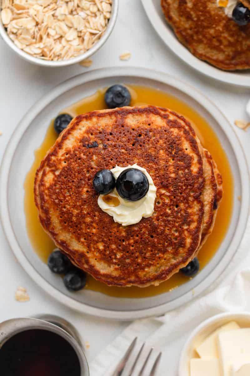 Stack of pancakes on a plate with blueberries, butter and syrup with more toppings around it. 