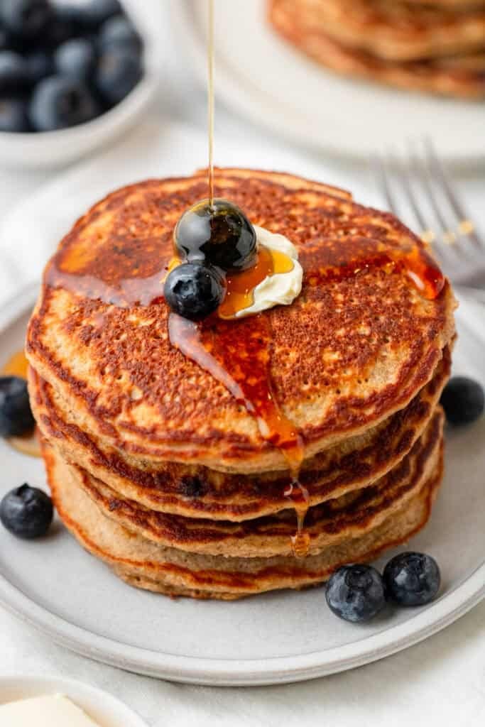 Cooked protein pancakes with blueberries stacked on a plate with syrup being drizzled on them with more pancakes behind it. 