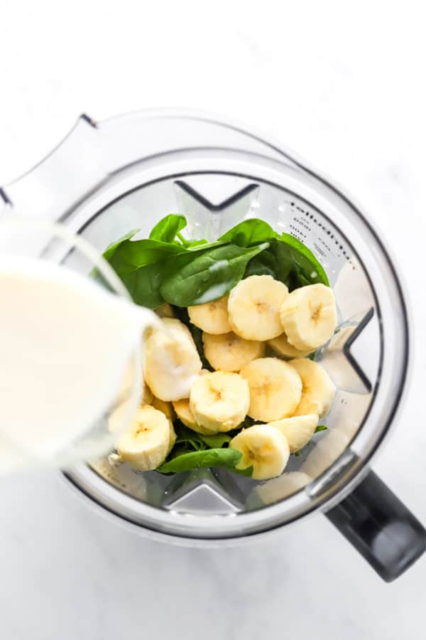 Pouring milk into a blender filled with spinach and sliced banana in a blender pitcher