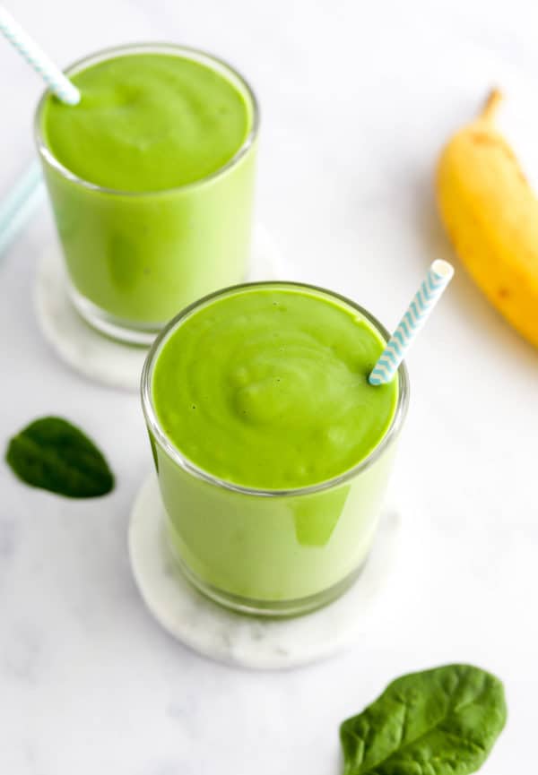 Two glasses of green drinks with straws in them with a whole banana next to them.