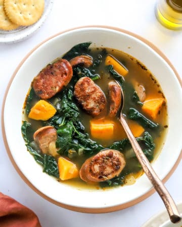 Soup with sliced sausage , kale and sweet potato in a round, white soup bowl with a spoon in the soup.