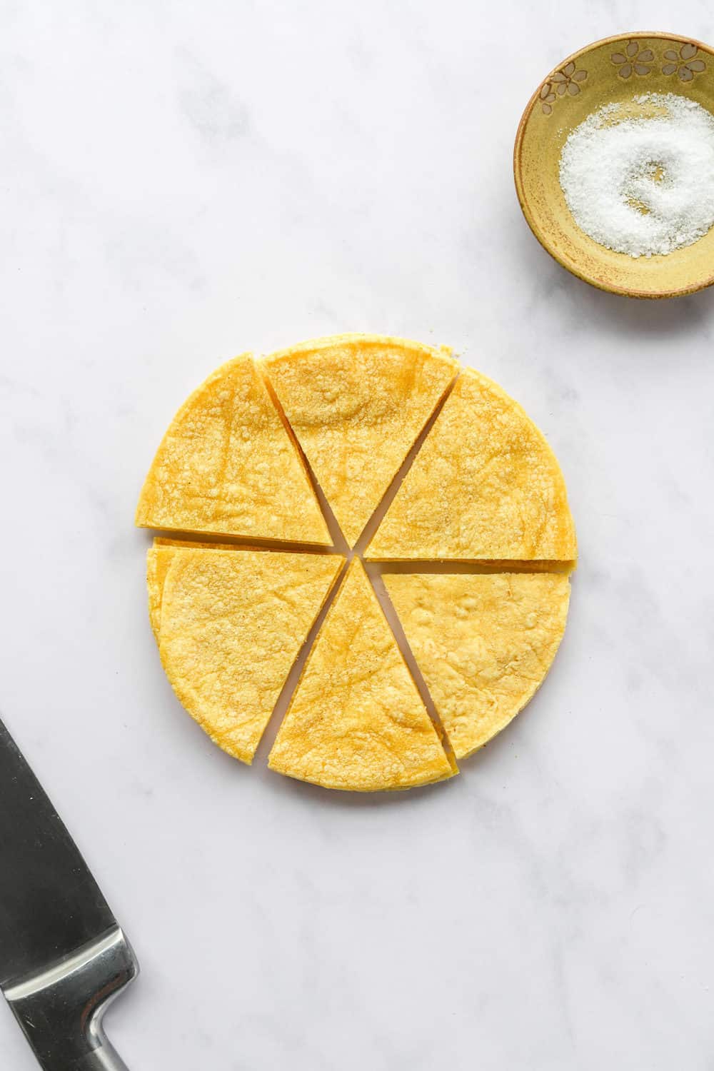 Stack of round yellow corn tortillas cut into 6 triangles with a knife next to the them and a yellow dish with salt in it behind them.