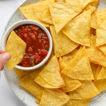 Hand dipping a tortilla chip in a white dish filled with tomato salsa.