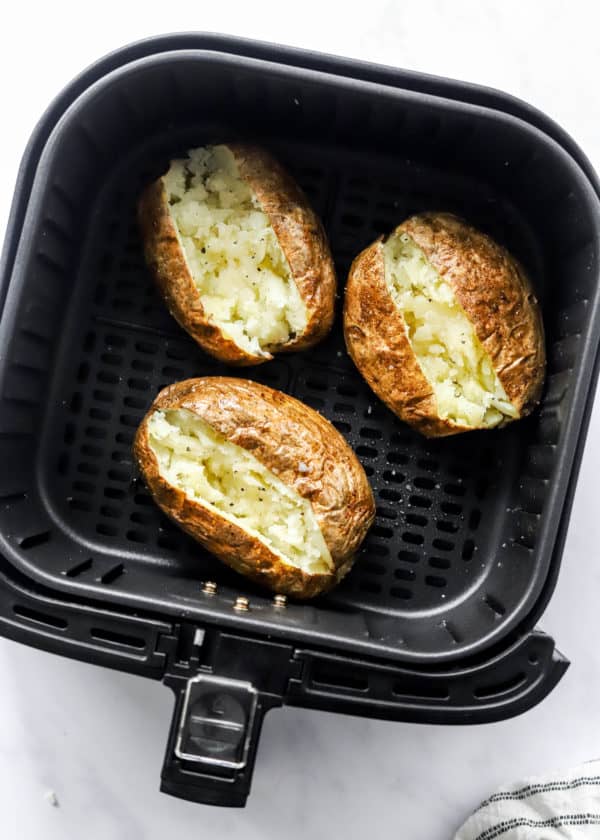 Fluffy cooked brown potatoes, slit open in a black, square air fryer basket.