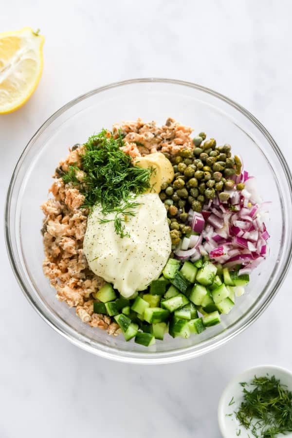 Pink salmon chunks a round glass bowl with capers, diced red onion, diced cucumber, mayo, dijon mustard and fresh dill.