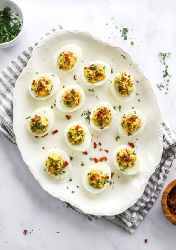 Oval platter filled with deviled eggs topped with chopped cooked bacon and herbs on top of a stripped towel.